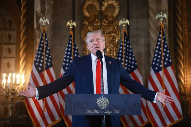 Donald Trump przemawia z podium z rękami wyciągniętymi przed czterema amerykańskimi flagami